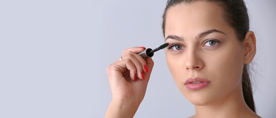 Young woman applying mascara against light background with space for text
