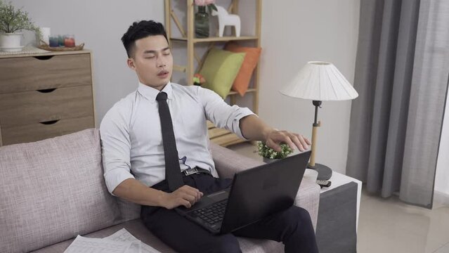 slow motion tired asian businessman done with work is relaxing by stretching arms and exercising neck after closing computer lid on living room couch at home.