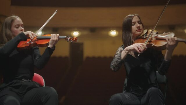 female violinists are playing music in orchestra, sitting on scene of philharmonic hall