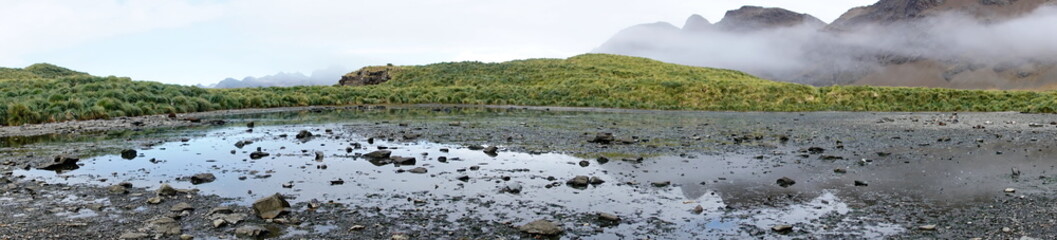 Panorama of a shallow, rocky inland lagoon below cloud shrouded mountains at Jason Harbor, South...