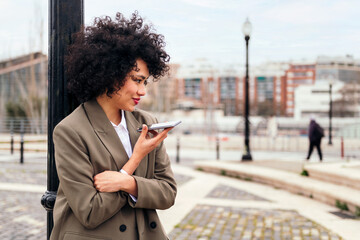 smiling young latin woman recording a voice message with her phone leaning against a city lamppost, technology and communication concept, copy space for text