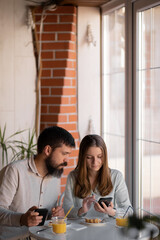 Portrait of young joyful couple using cell phones and laughing while having breakfast in home