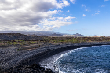 Scenic view on black stone pebble beach Playa Colmenares near Golf del Sur, Tenerife, Canary Islands, Spain, Europe. Mountain Teide, volcanic crater landscape covered by clouds. Waves Atlantic Ocean
