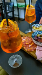 Typical italian aperitif (aperitivo) served with snacks at bar in Catania, Sicily, Italy, Europe. Aperol Spritz and slices of orange. Wooden plate with salami, ham (prosciutto), dried tomatoes, olives