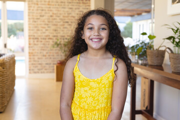 Portrait of smiling biracial cute girl with long curly hair wearing yellow dress standing at home - Powered by Adobe