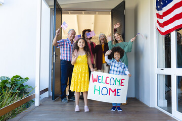 Multiracial cheerful multigeneration family with welcome home text and flags of america at entrance