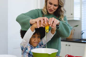 Multiracial mother making face while assisting son with afro hair in breaking egg in bowl in kitchen