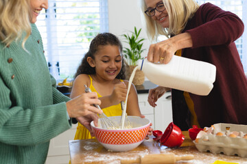Multiracial senior woman pouring milk while mother and daughter whisking batter in bowl in kitchen - obrazy, fototapety, plakaty