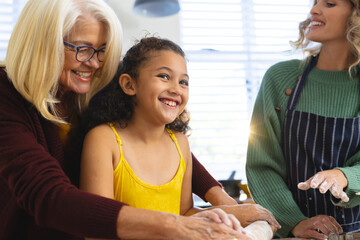 Happy woman looking at grandmother assisting granddaughter in rolling dough on table in kitchen
