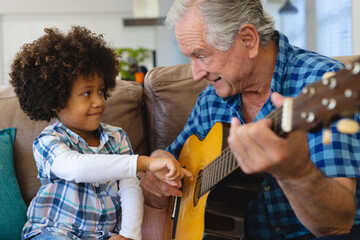 Multiracial grandfather holding grandson's hand and teaching him to play guitar on sofa at home