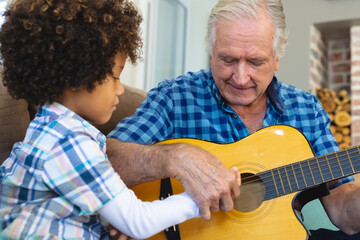 Caucasian senior man teaching playing guitar to biracial grandson while sitting at home - Powered by Adobe