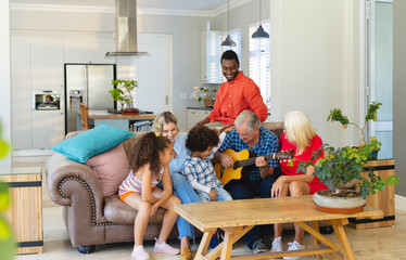 Multiracial senior man playing guitar while sitting with happy multigeneration family on sofa