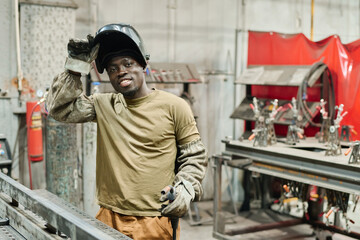 Portrait of African young welder in mask smiling at camera while working with metal products at...