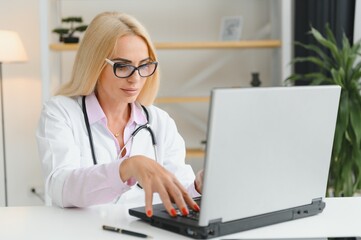 Portrait shot of middle aged female doctor sitting at desk and working in doctor office