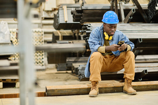 African young worker in hardhat scrolling in his smartphone during break at factory