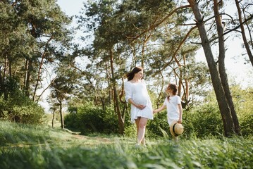 Happy family: a young beautiful pregnant woman with her little cute daughter on a sunny summer day. Parents and kids relationship. Nature in the country.