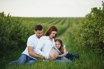 Fototapeta premium Beautiful pregnant brunette mom and dad and dauther in white clothes look at each other in nature.