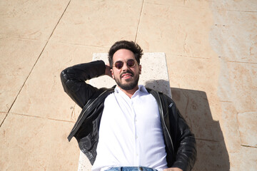 Obraz premium Portrait of handsome young man with beard, sunglasses, leather jacket and white shirt, lying on a stone bench. Concept beauty, fashion, trend, peace, relax, tranquility.