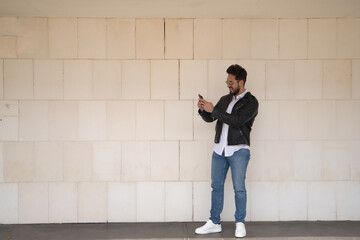 Fototapeta na wymiar Handsome young man with beard, sunglasses, leather jacket, white shirt and jeans, next to a white wall, consulting his cell phone. Concept beauty, fashion, trend, smartphone, app, social networks.