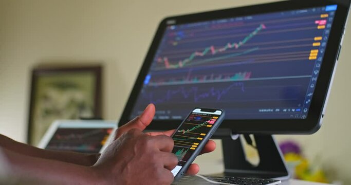 Man swiping with 2 fingers stock charts while day trading on his cellphone and his computer at his home office area working from home.