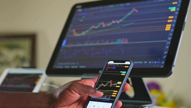 Man swiping with his thumb stock charts while day trading on his cellphone and his computer at his home office area working from home.