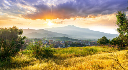 view from hill with golden grass and green bushes to a valley town with majectic mountains and scenic cloudy sunset on background