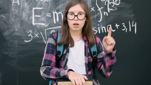 Teen girl little student in glasses and blue backpack standing and thinking on chalkboard background. Then she is pointing up with finger because finds answer. Brainstorming and Bright idea concept.