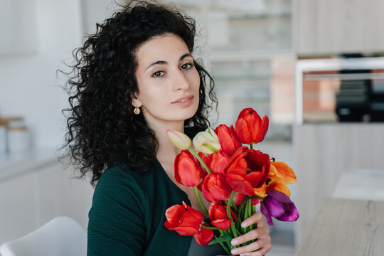 Pretty greatful curly italian young woman sitting at table holds colourful tulips looks at camera . Remote working hispanic businesswoman received flowers on women day, mother's day. Happy valentine.