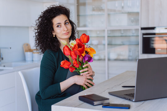 Confident greatful curly italian young woman sitting at table with laptop holding colourful tulips . Remote working hispanic businesswoman received flowers on women day, mother's day. Happy valentine.