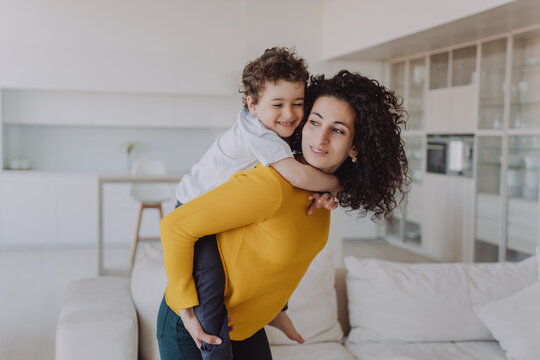 Young Spanish Curly Woman Having Fun With Son, Rolls Him On Back Smiling Wide. Babysitter In Yellow Sweater And Green Pants Entertains Little Cheerful Child At Home. Elder Sister Plays With Brother.