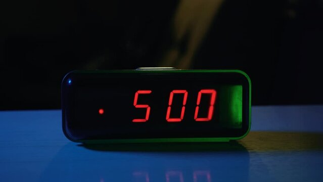 The electronic alarm clock on the table rings at 5:00 in the morning. The hand turns it off by pressing a button. Start of the day and wake up for work or study