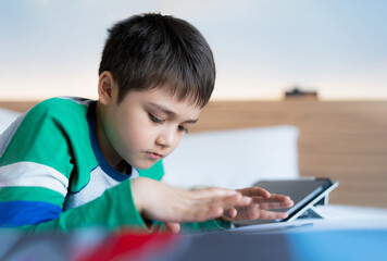 Cinematic portrait Kid playing games online on internet with friends in bed room, Young boy using tablet typing or chatting  at night, Childhood sitting on bed relaxing at home on weekend.