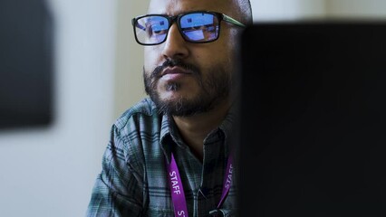 A close up shot from behind a work desk as an Asian office employee sits reading an email with blue light from his computer screen reflecting in his optical glasses