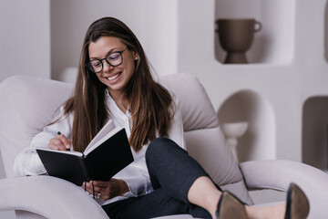 Satisfied brunette businesswoman in glasses, shirt black pants sitting on cozy chair with opened planner smiling broadly, checking to do list. Pretty hispanic young woman at home preparing for a day.