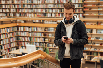 Fototapeta Smiling guy with a smartphone in a library. Student with headphones against bookshelves in library. obraz