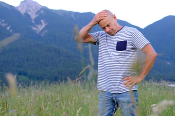 sad mature man, senior 60 years old standing in mountains above city, holds head with hand, Alps are behind him, green grass in meadow, concept of enjoy life in old age, travel, active lifestyle