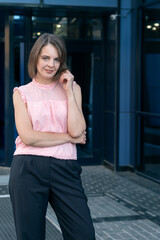 Fototapeta premium Portrait of woman in trousers and pink blouse against the backdrop of modern building. Successful business woman