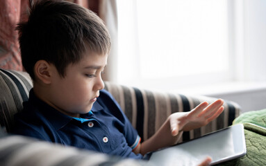 Fototapeta na wymiar Cinematic portrait young boy playing game on tablet sitting on sofa with light shining from window, Kid playing games online on internet at home, Child talking video call with friends at home
