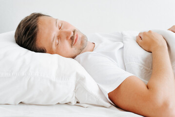 Fototapeta na wymiar Smiling sleeping man in bed, side view. Resting guy with closed eyes lying in bedroom indoors in early morning