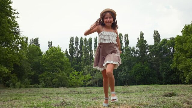 Happy Caucasian little girl in a straw hat and dress running skipping through the lawn in the park. Summer holidays. Slow motion. The concept of childhood and Children's Day.