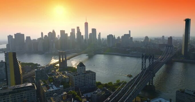 Manhattan and Brooklyn Bridges over East River at Golden hour. Hazy silhouettes of striking skyscrapers at backdrop of orange sky.