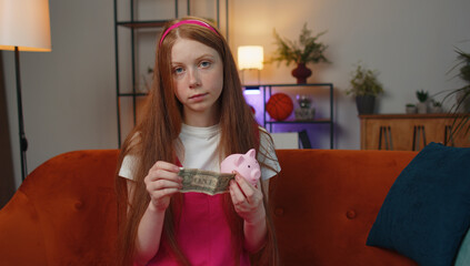 Poor young teen child girl insufficient amount of money, holding piggybank and one dollar banknote at home. Financial crisis. Bankruptcy. Poverty and destitution. Ginger kid in living room on couch