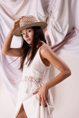 Portrait of beautiful woman in straw hat and white dress posing isolated over textile background. Romantic outfit.