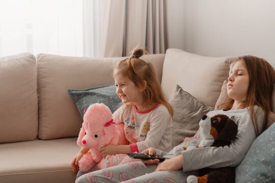 Two little girls sisters wear pyjamas sitting on beige couch at home with plush soft toys,  resting, having fun together