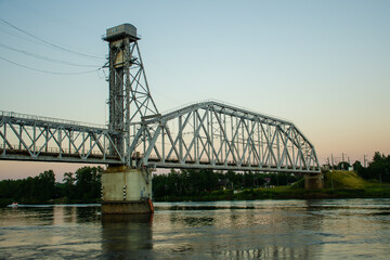 Modern metal bridge for transport across the river. The bridge is located outside the city, against the backdrop of sunset