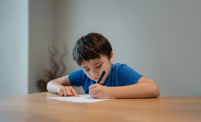 Naklejka premium Kid siting on table doing homework,Child boy holding black pen writing on white paper,Young boy practicing English words at home. Elementary school and home schooling, Distance Education concept
