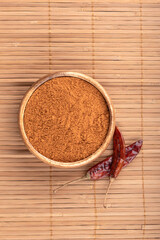 Wooden bowl with ground pepper and hot pepper pods on a food mat.