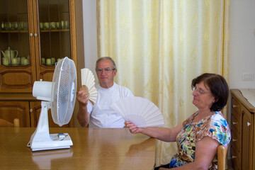 Image of an elderly couple at home wiping their sweat and trying to cool off with a fan and a fan....