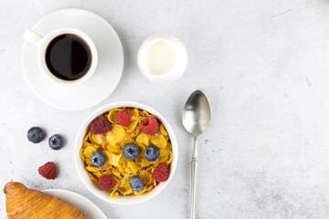 Obraz na płótnie Canvas Breakfast with corn flakes, berries: raspberries and blueberries, croissant, espresso and milk on a gray background. Top view, copy space.
