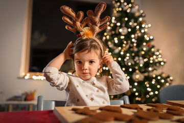 christmas, cooking and winter holidays concept - happy little baby girl with reindeer horns tiara...